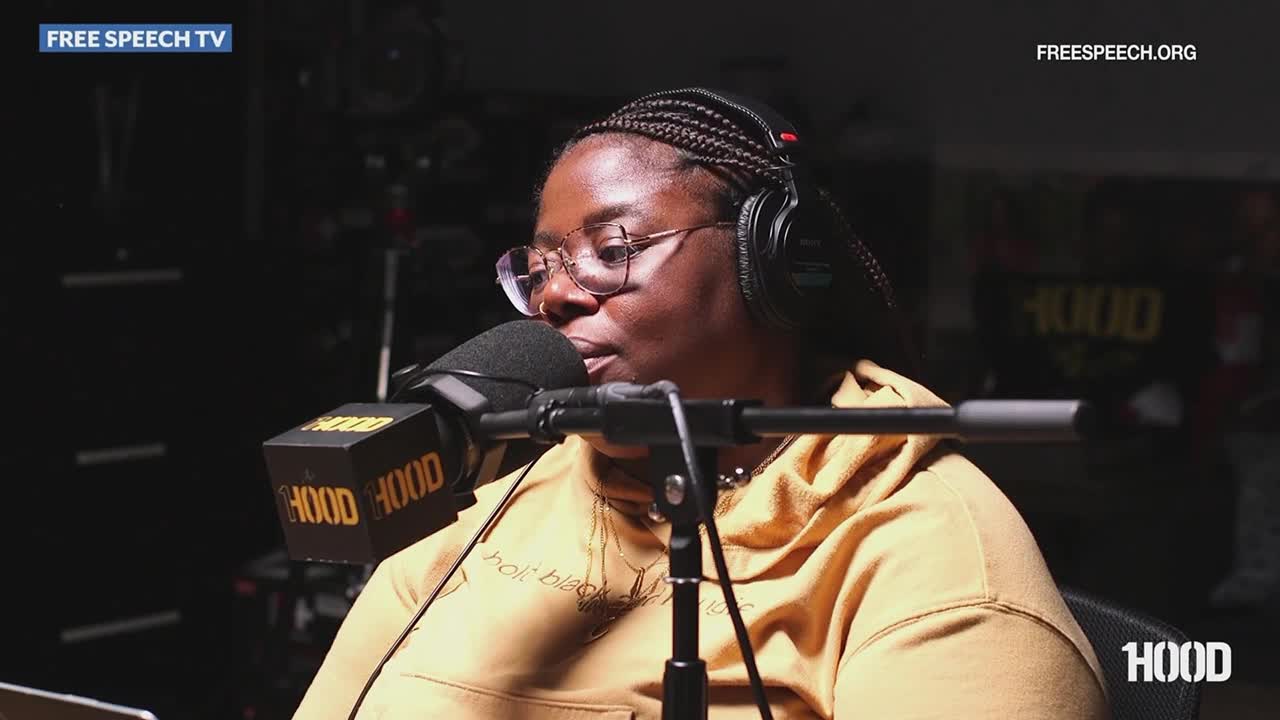 A woman with glasses and headphones speaks into a microphone, her face illuminated by studio lights.  The Free Speech TV logo is visible in the corner.
