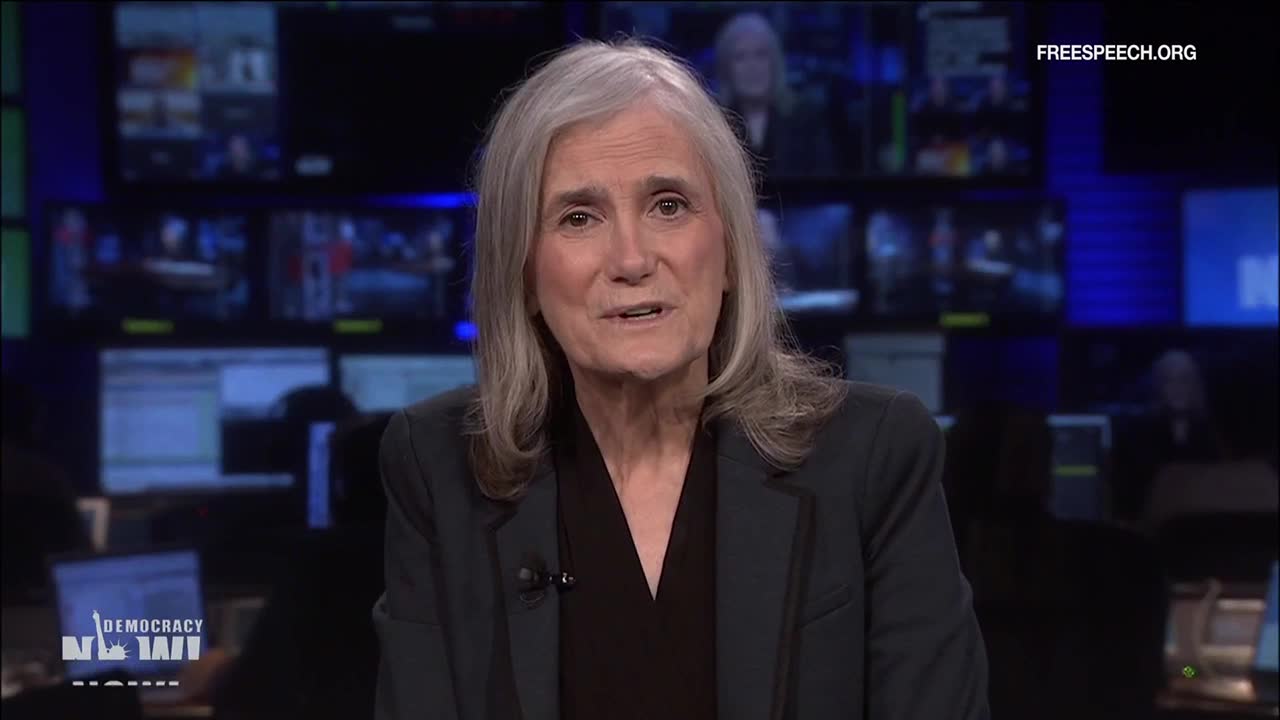 The woman with silver hair is speaking, her gaze directed toward the camera. Behind her, multiple screens display what looks like a newsroom, the Free Speech TV logo visible in the corner.

