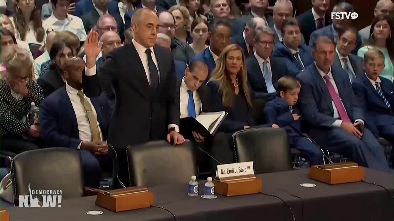A man in a dark suit raises his right hand, taking an oath before a panel. The scene is being broadcast by Free Speech TV, with a "Democracy Now!" logo visible in the corner.
