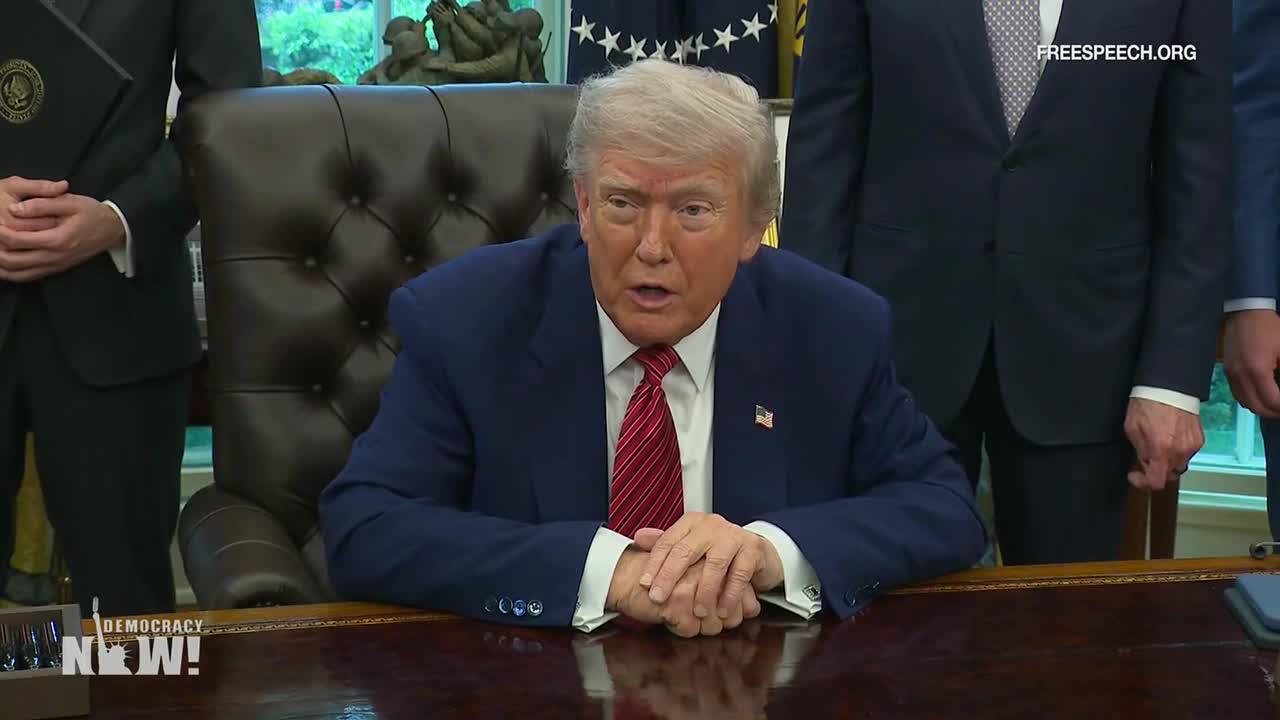 The former U.S. president sits at a large desk, speaking, his hands resting on the surface. Behind him, several men in suits stand silently, the scene broadcast on Free Speech TV.
