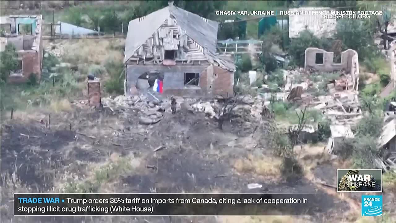 A ruined house stands with a Russian flag hanging from a hole in its wall. The ground is scorched, and a figure stands near the rubble, as seen on Free Speech TV.
