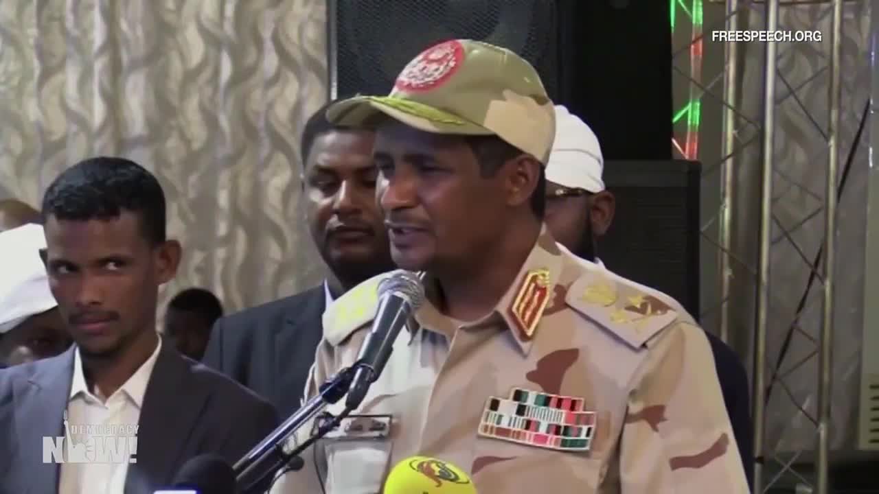 A man in a military uniform speaks into a microphone, his words broadcast by Free Speech TV. Behind him, a few other men stand, listening intently.
