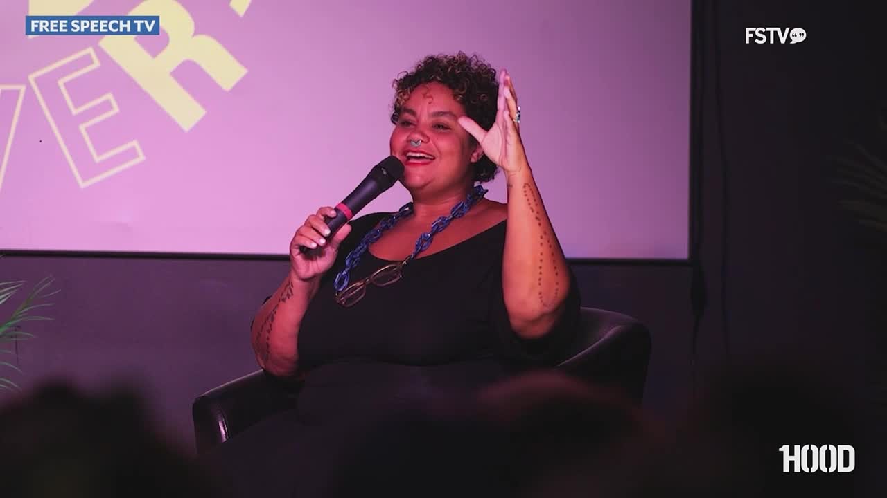 A woman sits in a chair, smiling and gesturing with her right hand while holding a microphone. The Free Speech TV logo is visible in the corner of the screen.
