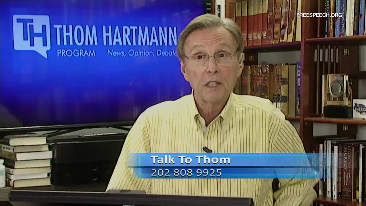 Thom Hartmann, wearing a yellow striped shirt, is speaking directly to the camera. Behind him, a screen displays the logo for "The Thom Hartmann Program" from Free Speech TV, and a phone number is visible at the bottom.
