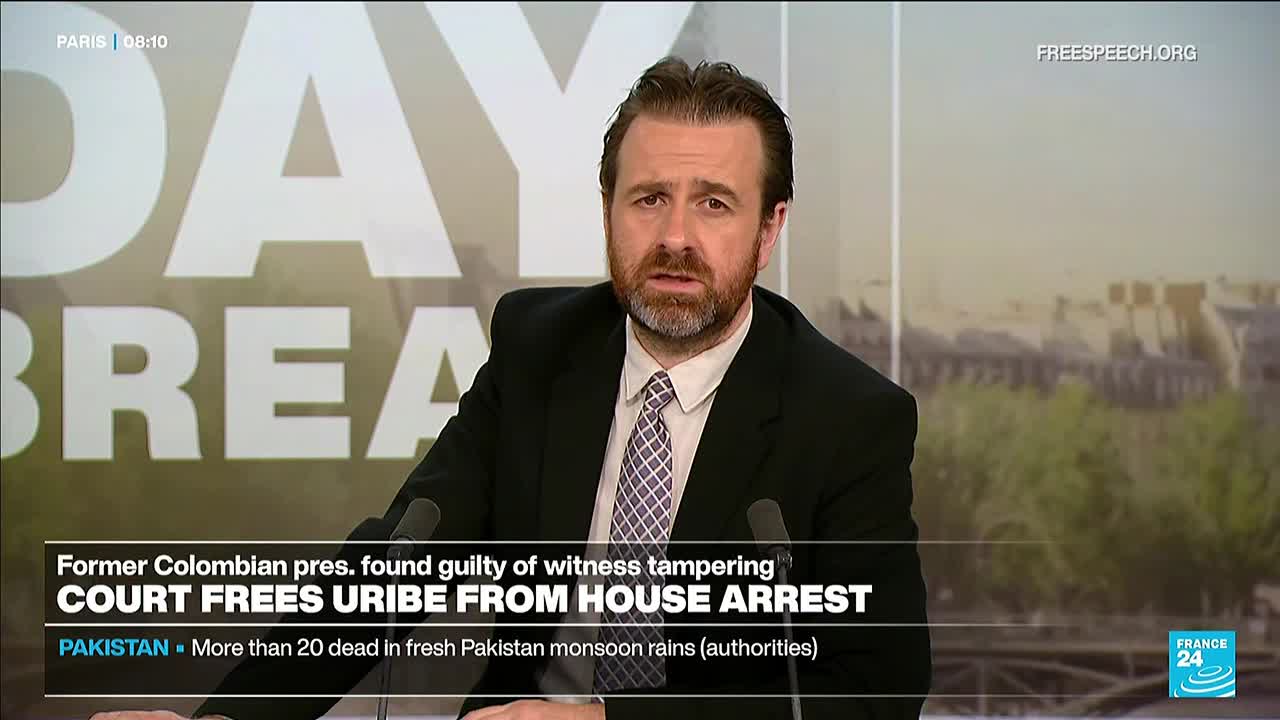 A man in a suit and tie is speaking into two microphones, his expression serious. Below him, the screen displays breaking news headlines from France 24, including a story about a Colombian president.
