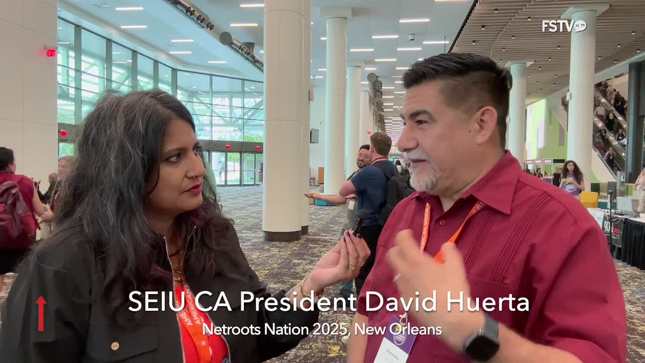 A woman with long, dark hair is holding a microphone, interviewing a man in a red shirt. He gestures with his hands as he speaks, presumably at the Netroots Nation conference, captured by Free Speech TV.
