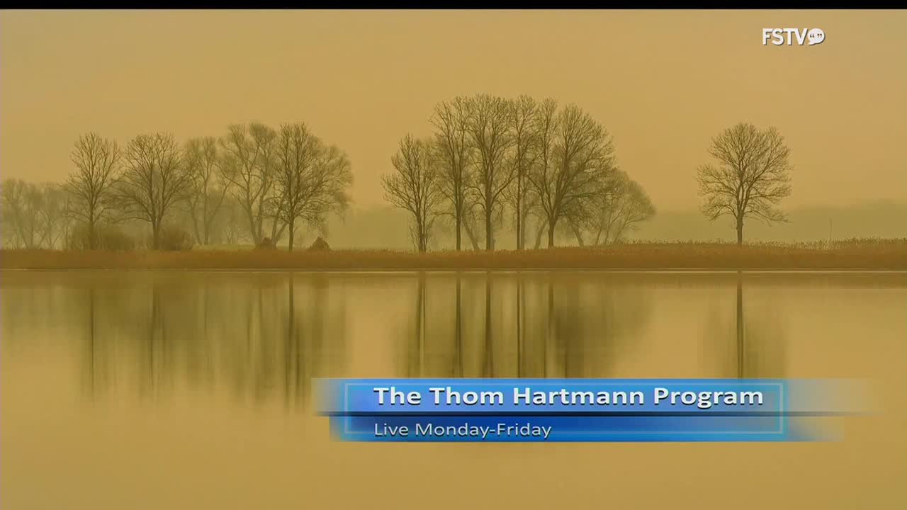 The image shows a tranquil lake with bare trees reflected in the water. A logo for Free Speech TV, FSTV, is in the upper right corner, and a lower third identifies "The Thom Hartmann Program."
