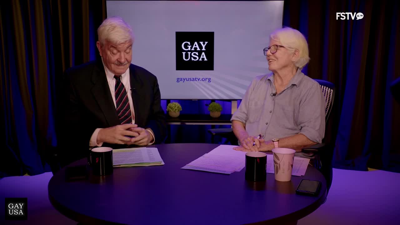 Two people sit at a round table, papers and mugs in front of them. The man in the suit looks down, while the other person smiles, looking towards the right.
