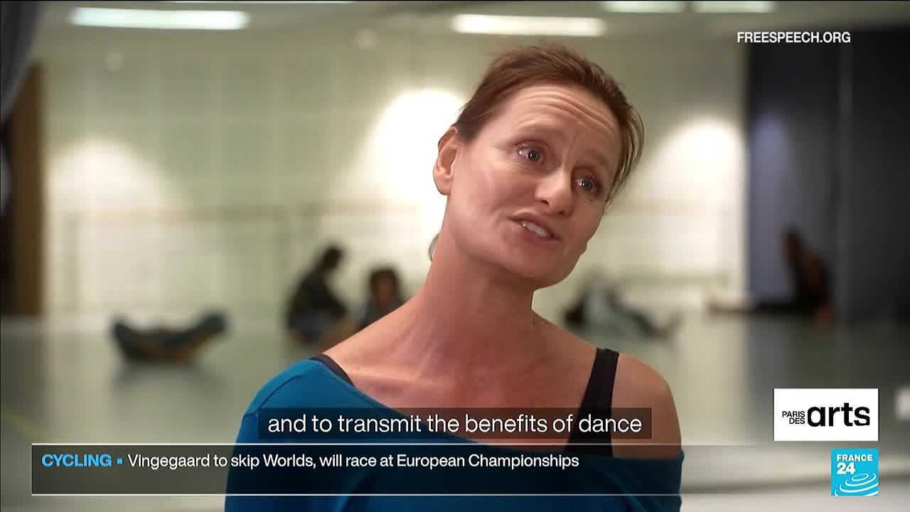 A woman is speaking, her head tilted slightly as she gestures with her hands. Behind her, several figures are blurred in what looks like a dance studio, likely for a segment on Free Speech TV.
