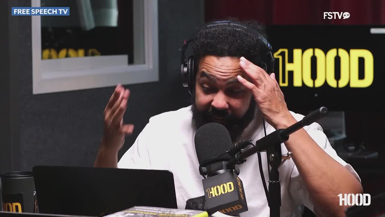 The man, wearing headphones, gestures with both hands as he speaks into a microphone. He's on a set for "HOOD" on Free Speech TV, with the show's logo glowing behind him.
