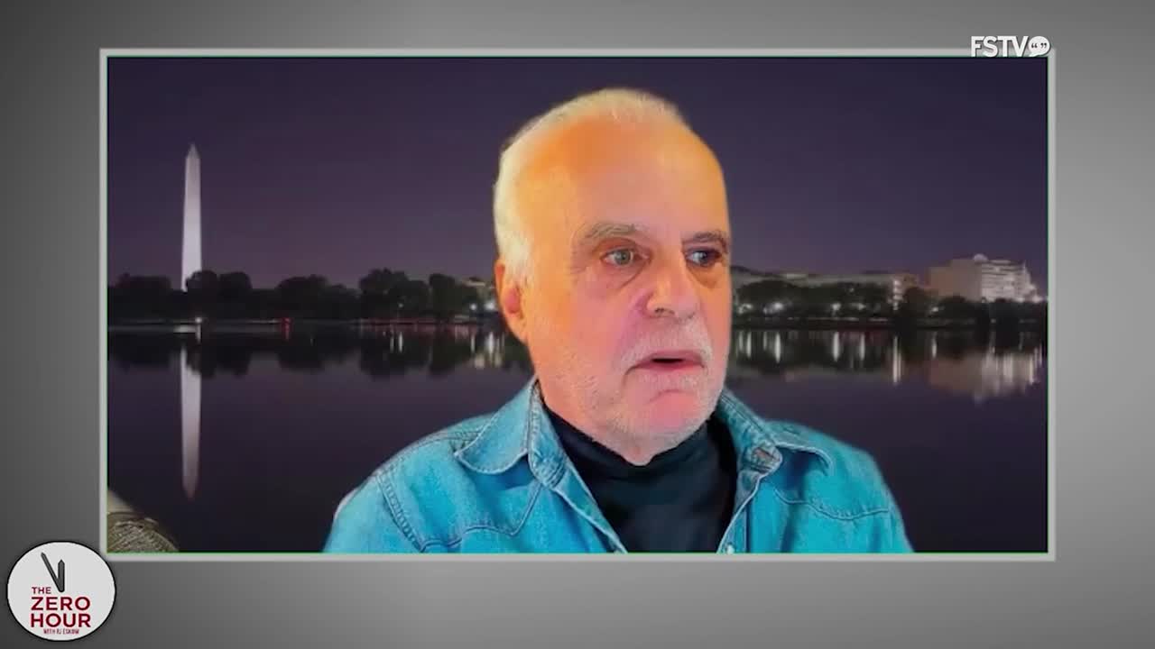 A man in a denim shirt and black turtleneck is centered on the screen, speaking. Behind him, a nighttime backdrop of the Washington Monument and reflecting pool is visible.
