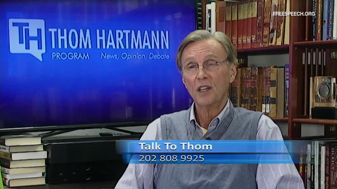 Thom Hartmann, wearing a sweater vest, is speaking directly to the camera, his mouth moving as he talks. Behind him, a screen displays the "Thom Hartmann Program" logo, and a lower third invites viewers to "Talk to Thom."
