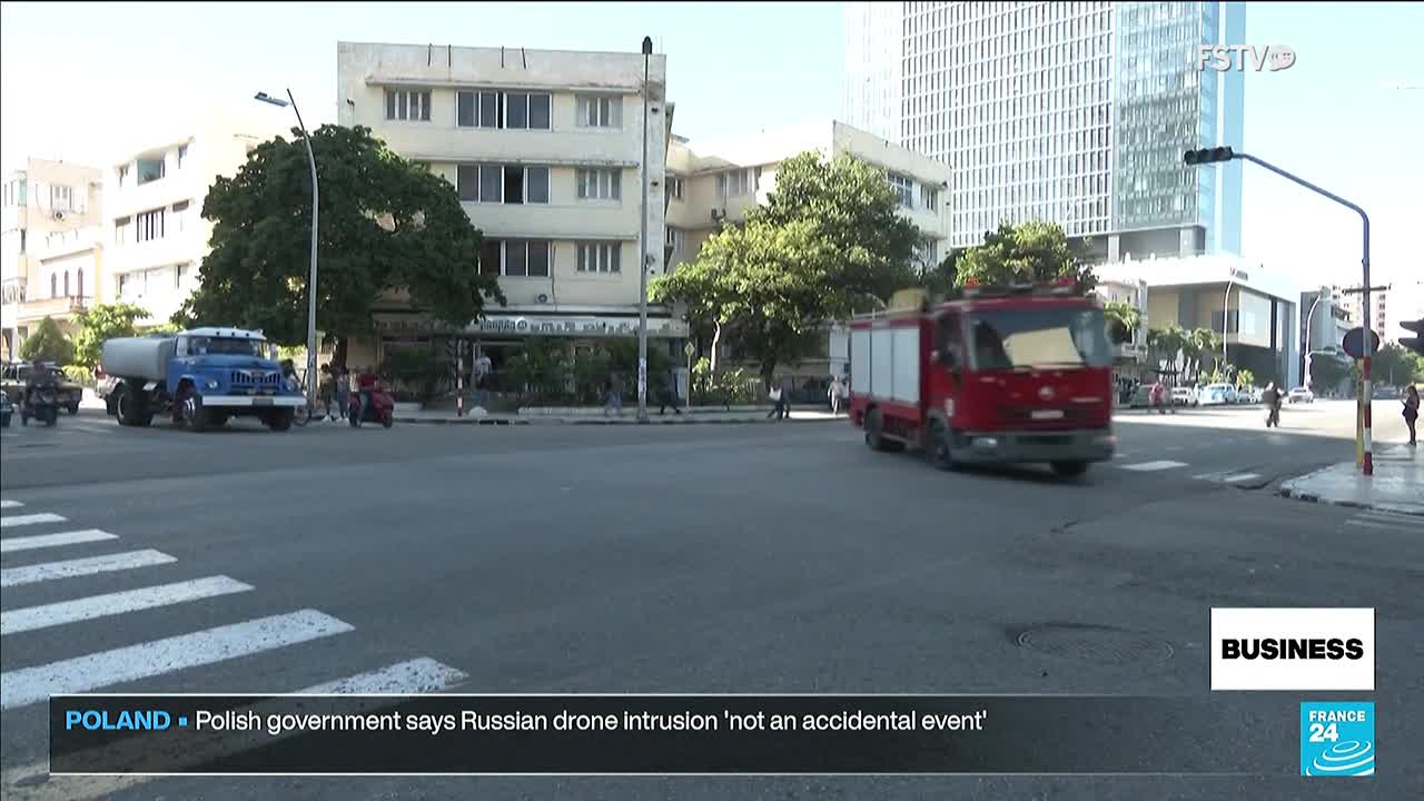A red fire truck speeds across the intersection, its lights off. A blue water tanker truck and other vehicles are also moving through the city streets.
