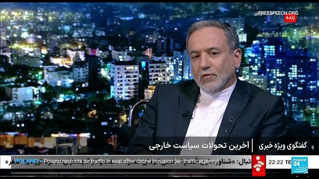 A man with a gray beard is seated, speaking on a Free Speech TV program. Behind him, a cityscape glows with lights against a dark sky.
