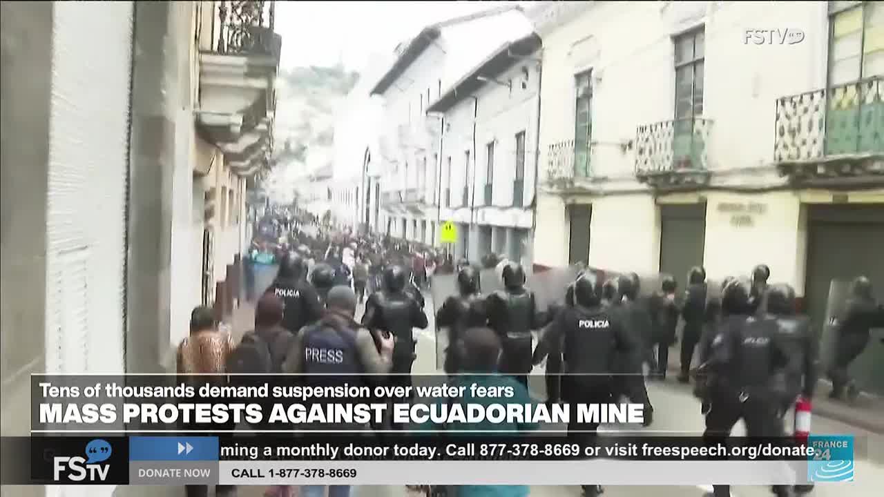 A line of police in riot gear moves down the street, pushing against a crowd of people.  The scene is being broadcast on Free Speech TV, with a banner describing mass protests against an Ecuadorian mine.

