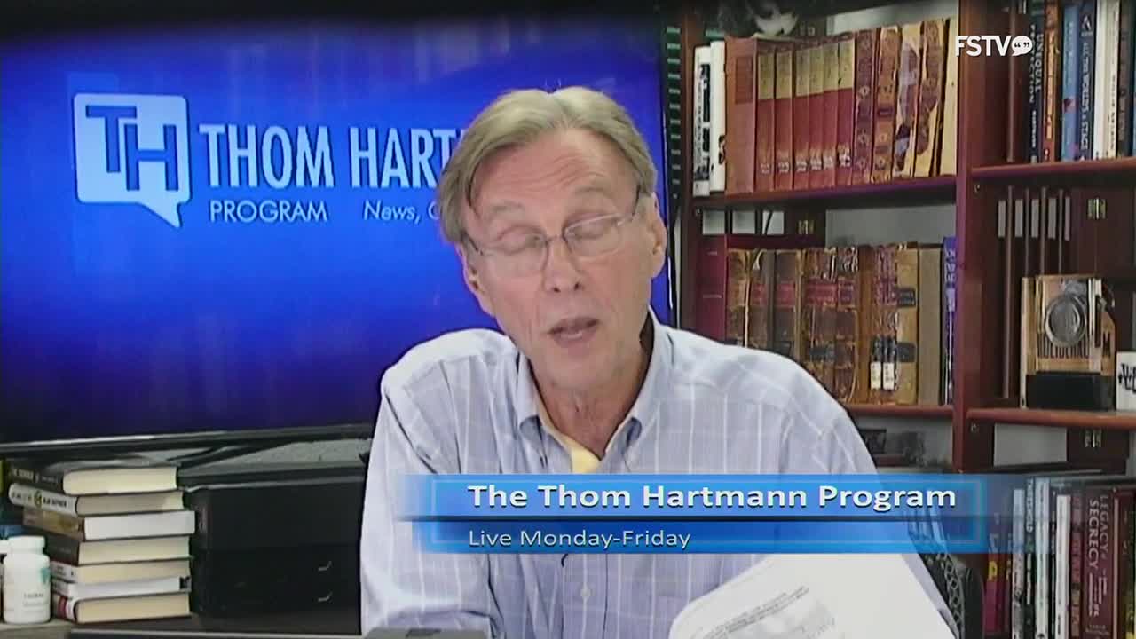 The host of "The Thom Hartmann Program" on Free Speech TV is speaking, reading from a paper. Behind him, a bookshelf is filled with books, and a screen displays the show's logo.
