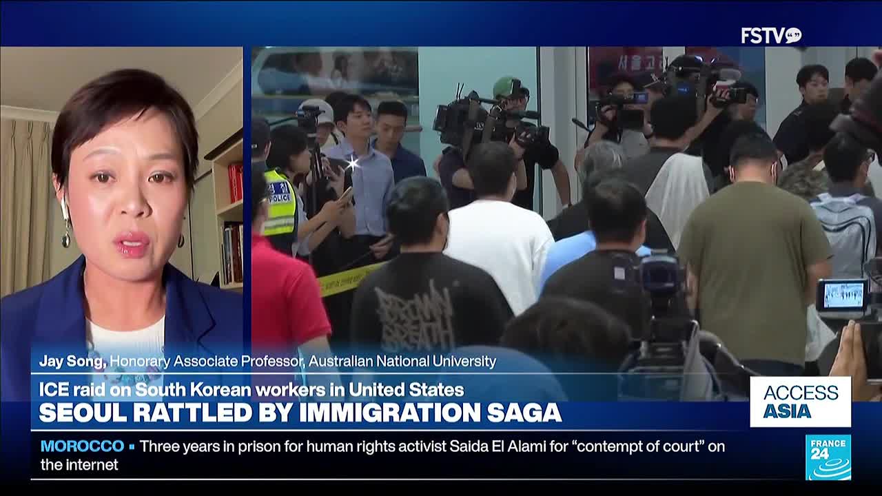 A woman on Free Speech TV discusses an ICE raid on South Korean workers. Behind her, a crowd gathers near a building, with police and media present.
