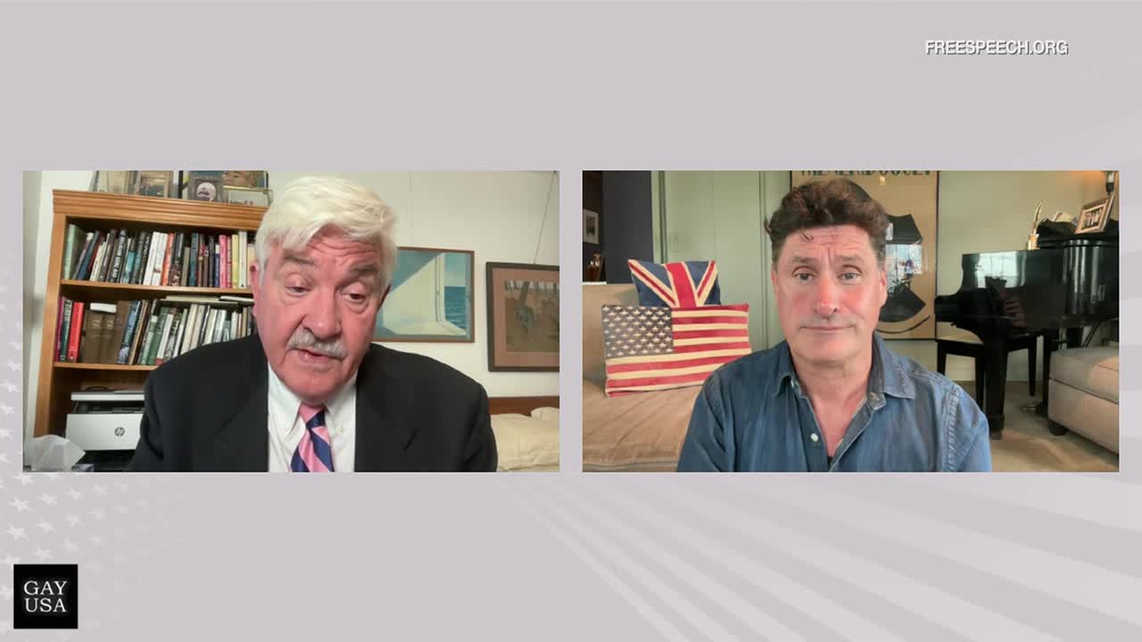 A man in a suit speaks intently, while another, in a denim shirt, listens from a room decorated with flags and a piano. This broadcast, from Free Speech TV, is also branded with "GAY USA."
