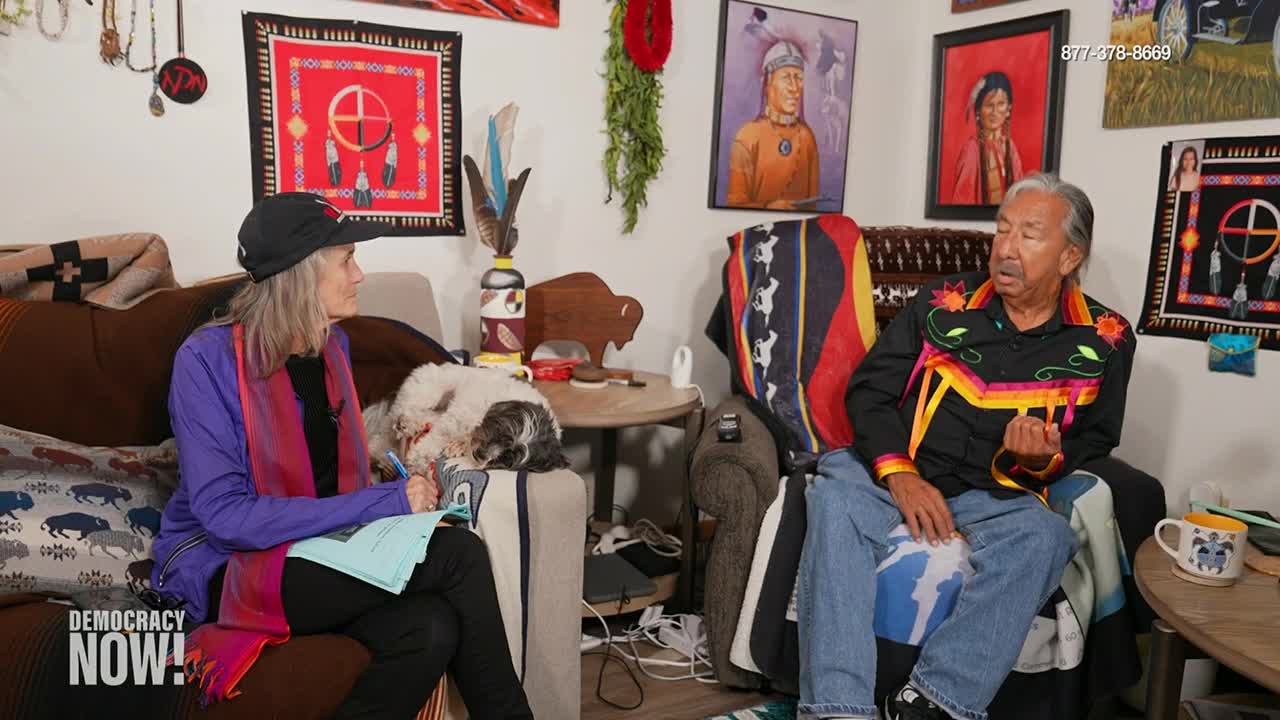 A man in a decorated shirt gestures as he speaks, seated on a couch. Across from him, a woman in a cap and scarf listens, pen in hand, as a small dog rests between them.
