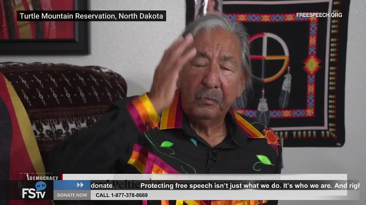 A man in a colorful shirt raises his hand to his forehead, eyes closed.  The Free Speech TV logo and a call to donate are visible at the bottom of the screen.
