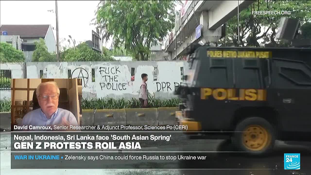 A black police vehicle with yellow lettering and wheels drives past a wall scrawled with "FUCK THE POLICE." A man in a suit is speaking on a screen, while a young person walks along the wall.
