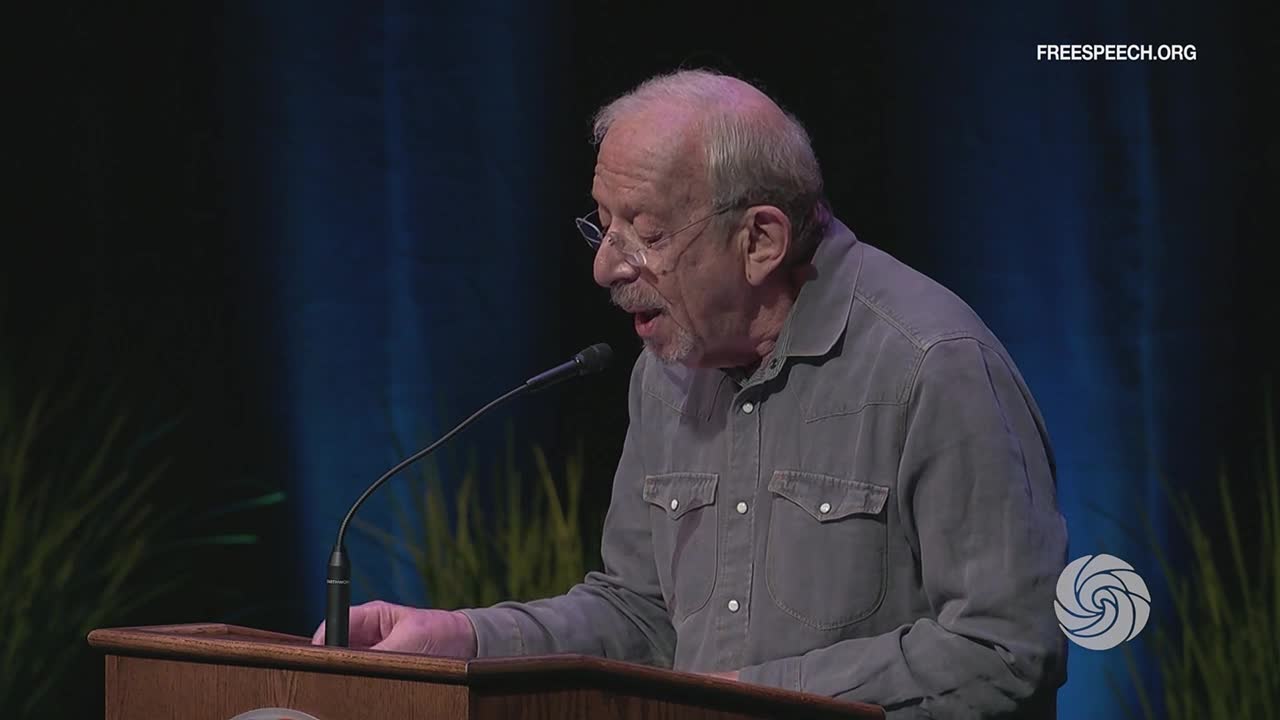A man in a gray shirt, wearing glasses, speaks into a microphone at a podium, likely for Free Speech TV. The backdrop is a dark blue with some green, and the FreeSpeech.org logo is in the corner.
