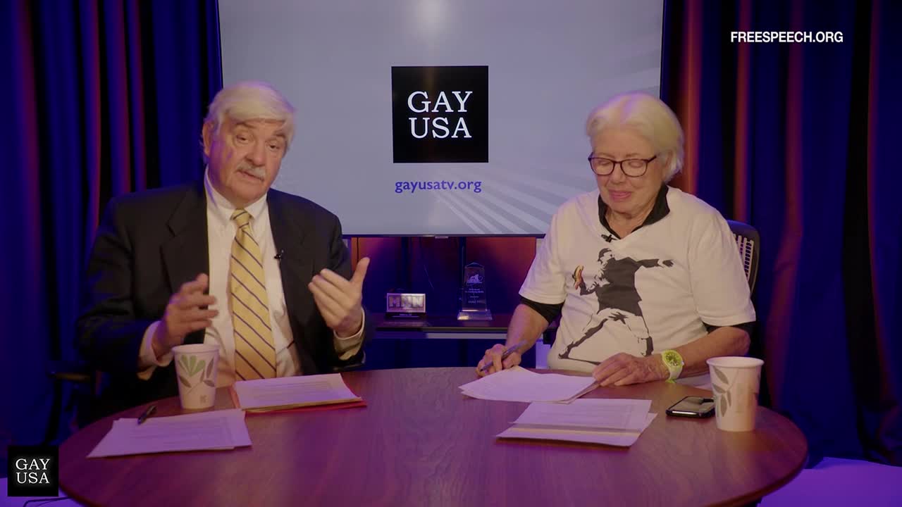 A man in a suit gestures with his hands as he speaks, while a woman beside him, wearing a Banksy t-shirt, looks down at papers on the table. The "GAY USA" logo is visible behind them, and the Free Speech TV logo is in the top right corner.
