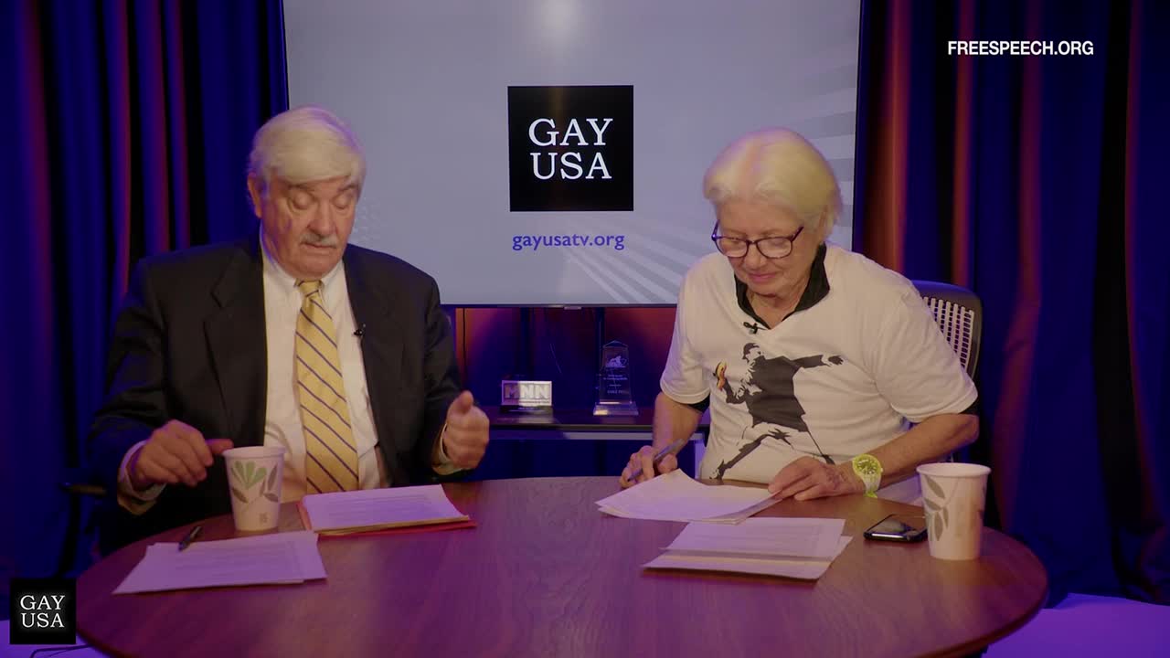 Two people sit at a round table, papers in front of them, as if they're about to discuss something on Free Speech TV. The man in the suit gestures with his hands while the woman, wearing a Banksy t-shirt, looks down at her notes.
