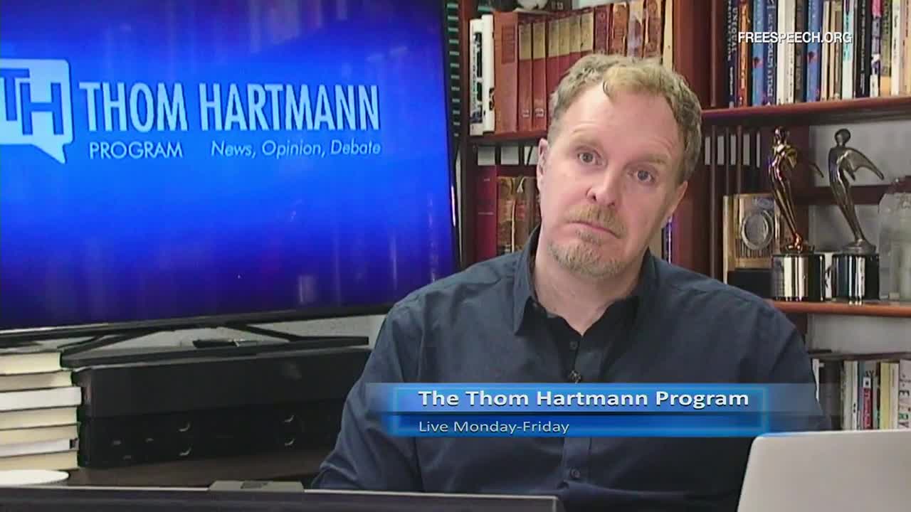 A man with reddish hair and a beard looks directly at the camera, his expression serious. Behind him, a large screen displays the logo for "The Thom Hartmann Program" on Free Speech TV.
