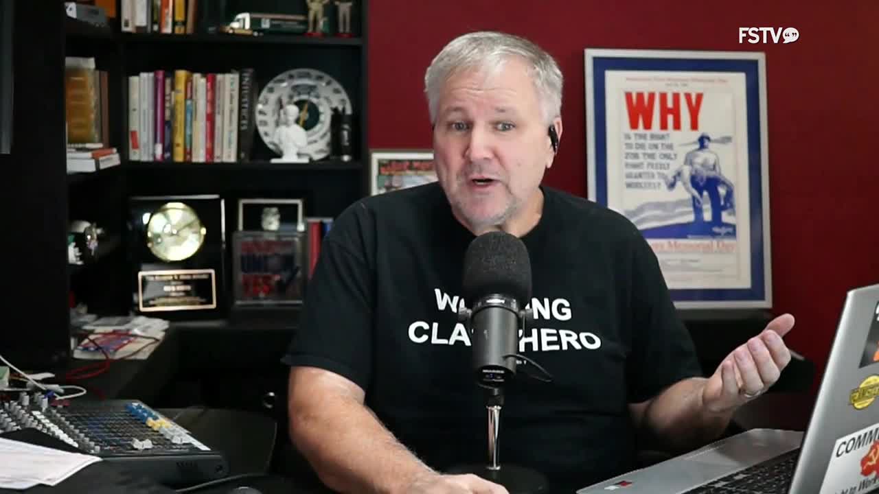 A man in a black t-shirt speaks into a microphone, gesturing with his right hand. Behind him, a "Free Speech TV" logo is visible, along with a shelf of books and a framed poster.
