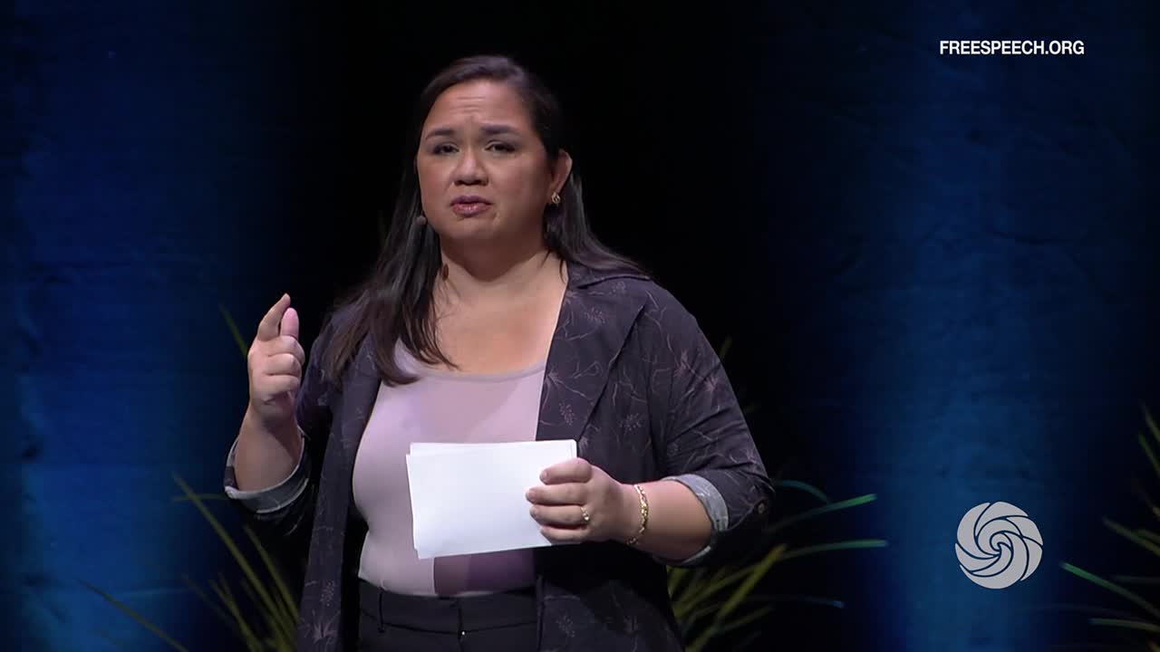A woman, holding a stack of papers, gestures with her right hand as she speaks. The Free Speech TV logo is visible in the corner.
