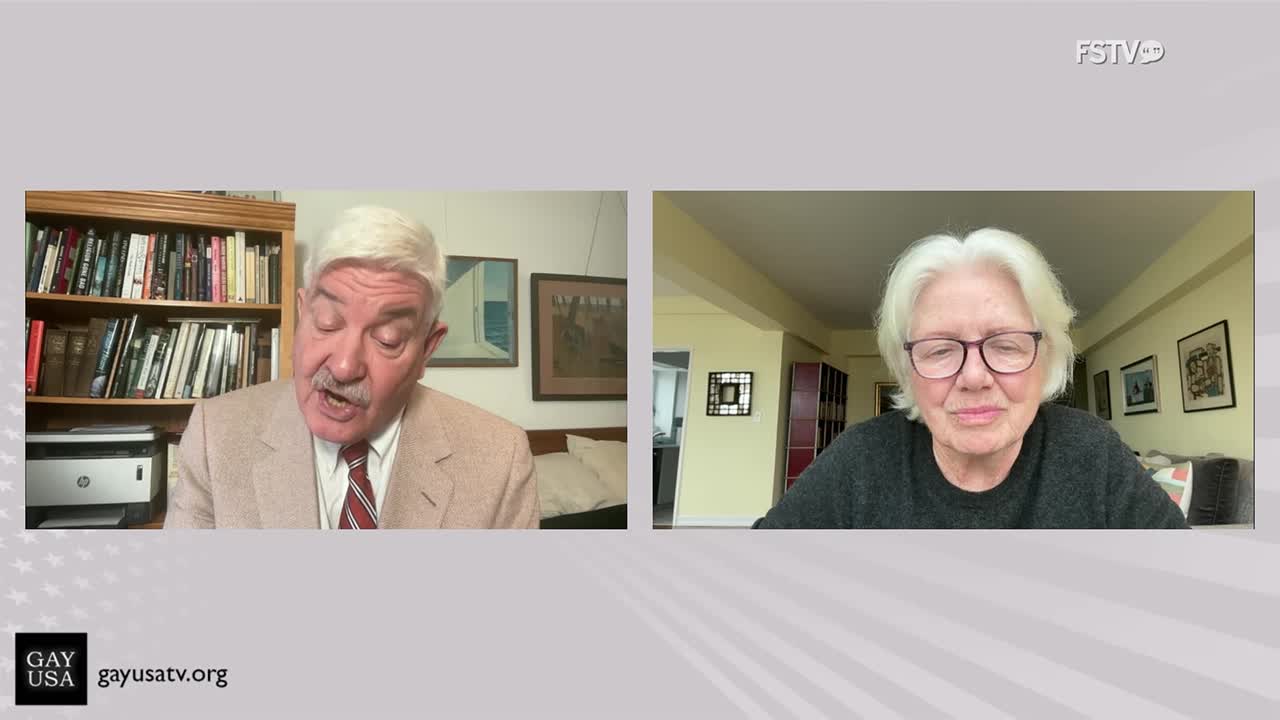 On Free Speech TV, a man in a beige blazer speaks animatedly from a room with a bookshelf. Across the screen, a woman with white hair and glasses listens intently.
