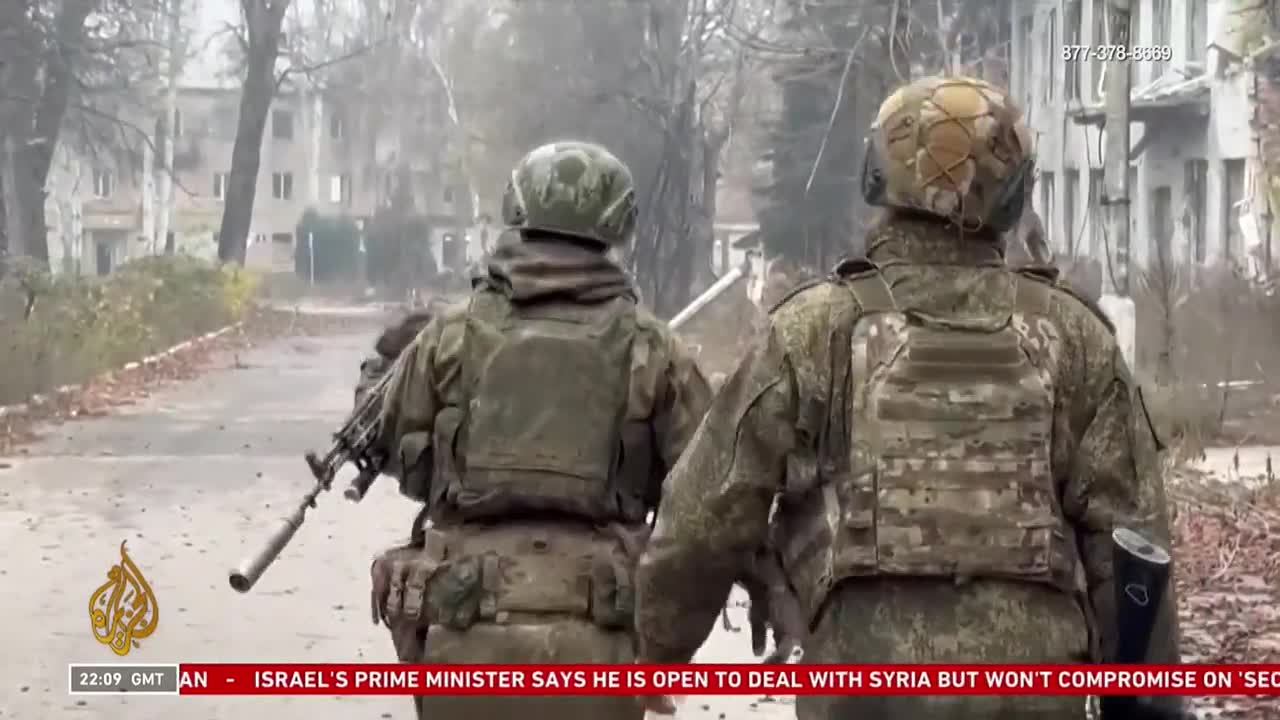 Two soldiers in camouflage gear walk down a debris-strewn street, rifles at the ready. Buildings with broken windows stand in the background, the scene broadcast on Free Speech TV.
