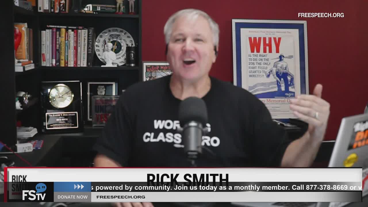 Rick Smith, on Free Speech TV, gestures with his right hand while speaking into a microphone. Behind him, a shelf is filled with books and memorabilia, and a poster reads "WHY."
