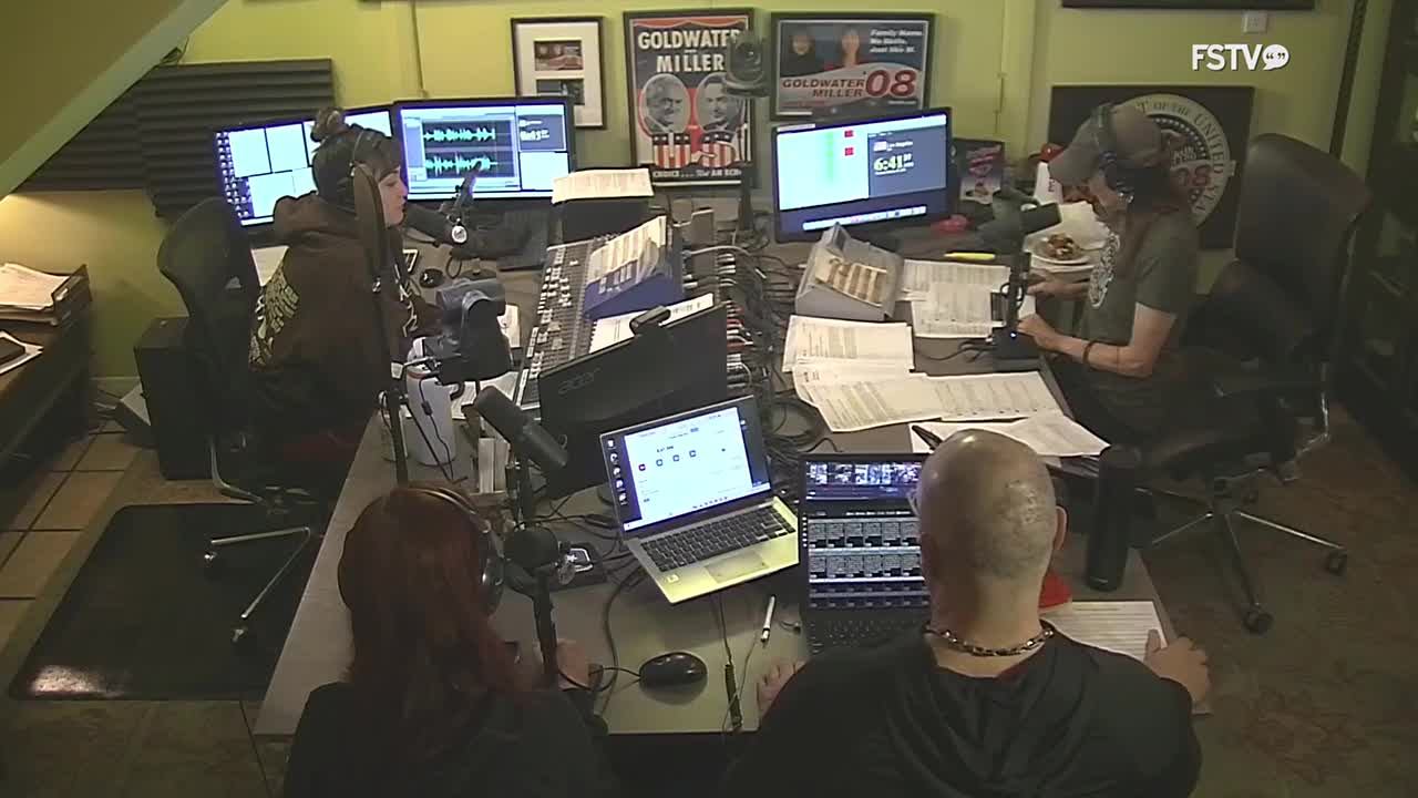 A woman with her hair in a bun leans into a microphone, headphones on, at a desk filled with audio equipment. Across from her, a bald man with a beaded necklace also wears headphones, focused on a laptop screen displaying audio levels. The FSTV logo is visible on a monitor in the background.