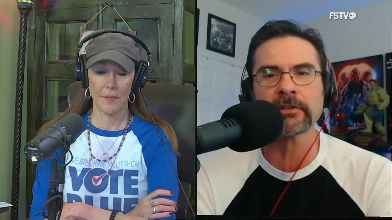 A woman wearing a "Vote Blue" shirt and headphones speaks into a microphone. To her right, a man with glasses and a goatee also speaks into a microphone, with comic book posters behind him.