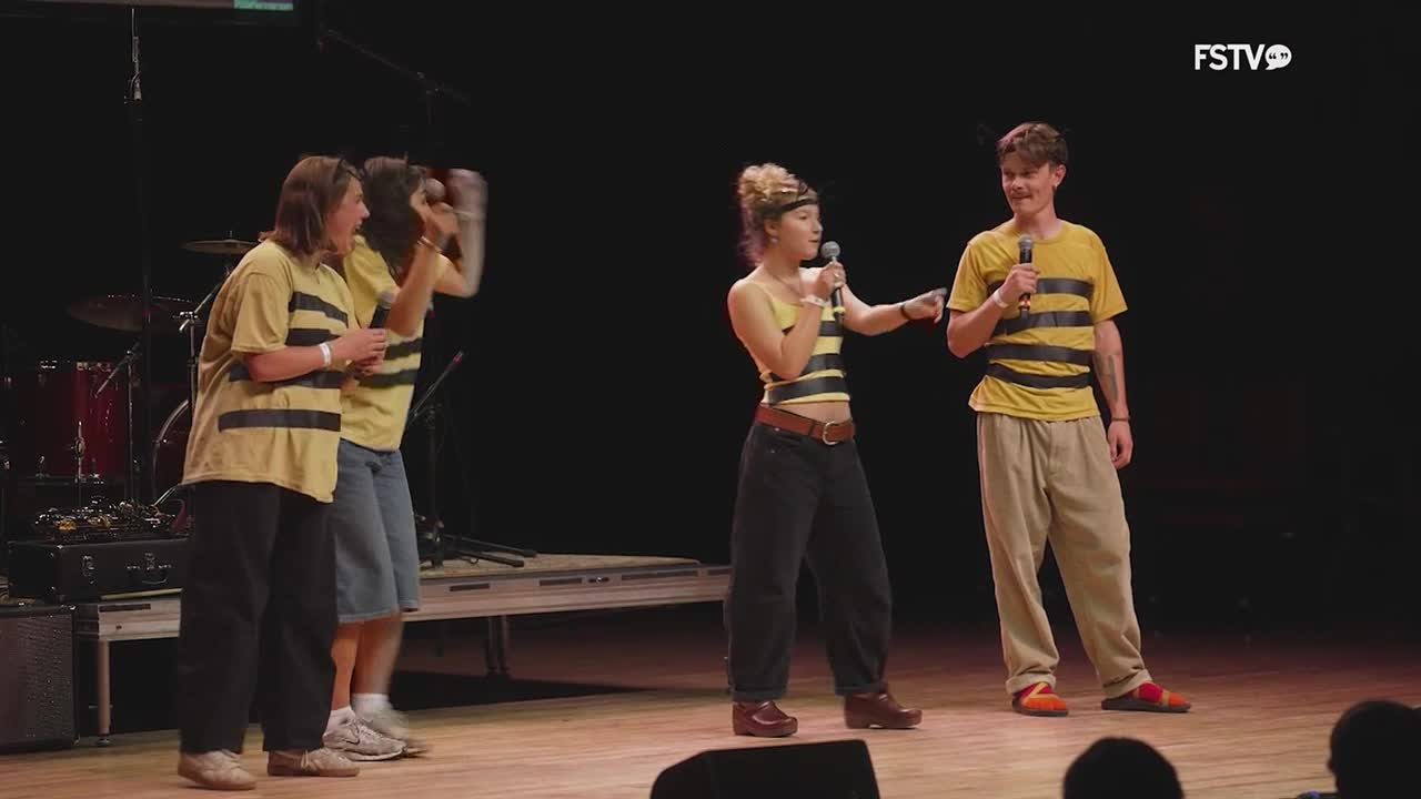 A group of young people in matching bumblebee-striped shirts are on stage, holding microphones. One woman points at a man as they both speak into their mics.