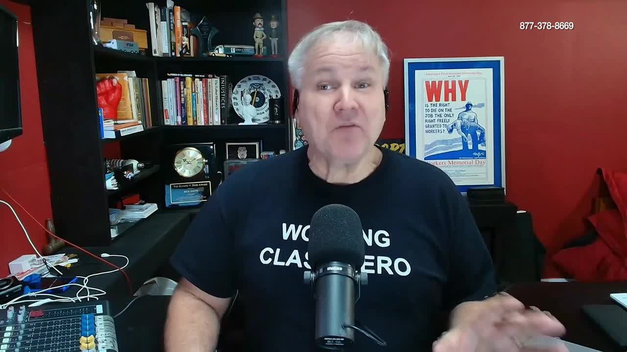 A man speaks into a microphone, his t-shirt reads "Working Class Hero." Behind him, shelves are packed with books and framed memorabilia, including a poster that says "WHY."