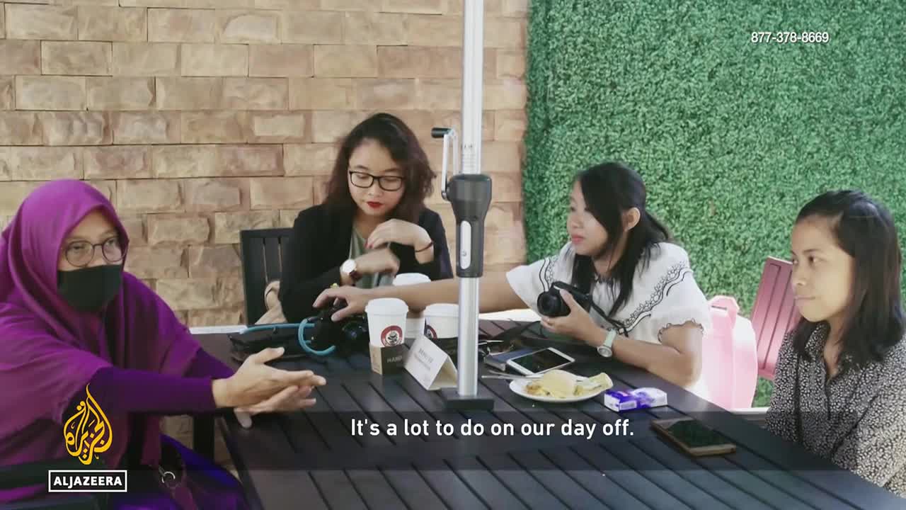 Four women sit at a table outdoors, with one in a purple hijab and face mask speaking animatedly. Another woman, wearing glasses and a dark blazer, reaches for a camera on the table.