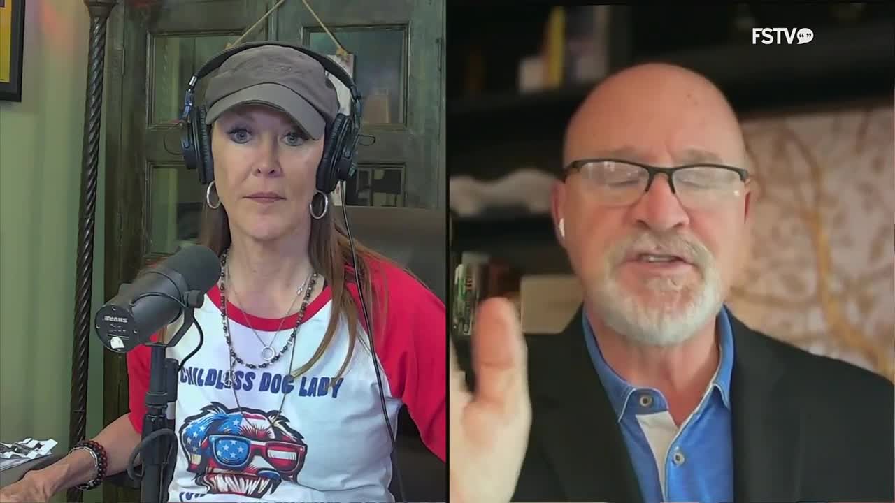 A man with glasses and a beard speaks, gesturing with his right hand. Across from him, a woman wearing headphones and a baseball cap listens intently.