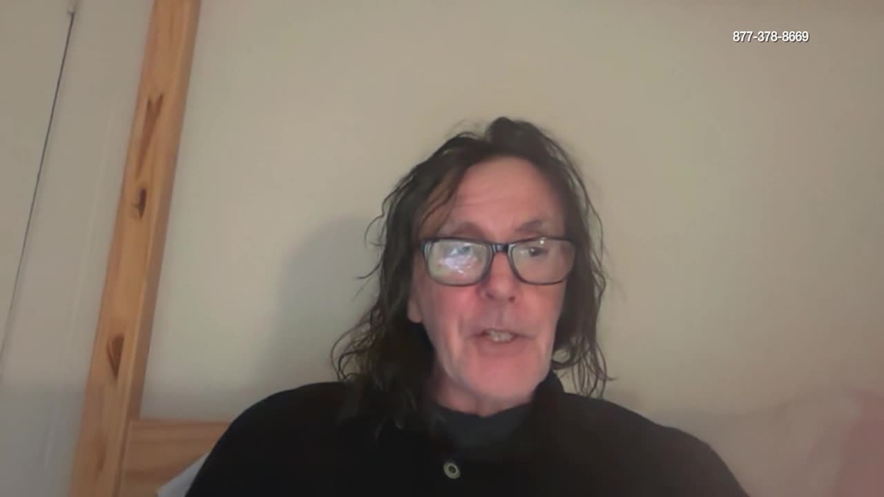 A man with long, dark hair and glasses speaks directly to the camera. A phone number appears in the upper right corner of the screen.