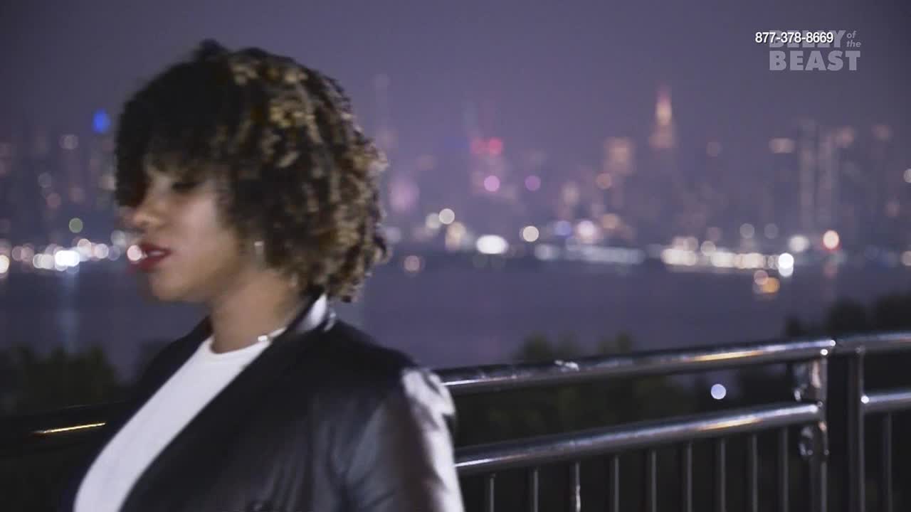 A woman with curly hair stands by a railing, looking out at a city skyline at night. The lights of buildings and cars twinkle across the water.