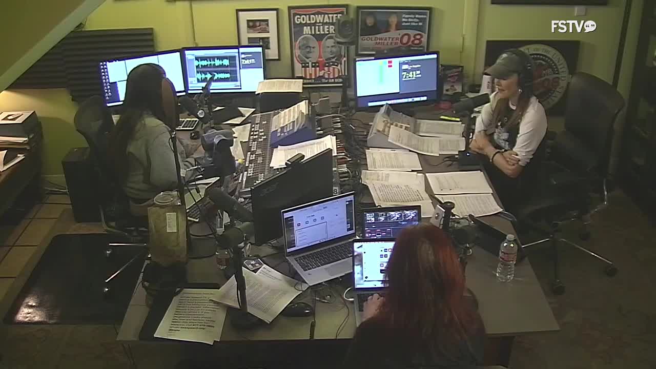 Two women are in a studio, one with headphones on, speaking into a microphone. Papers are spread across the desk, with multiple computer monitors displaying waveforms and text.
