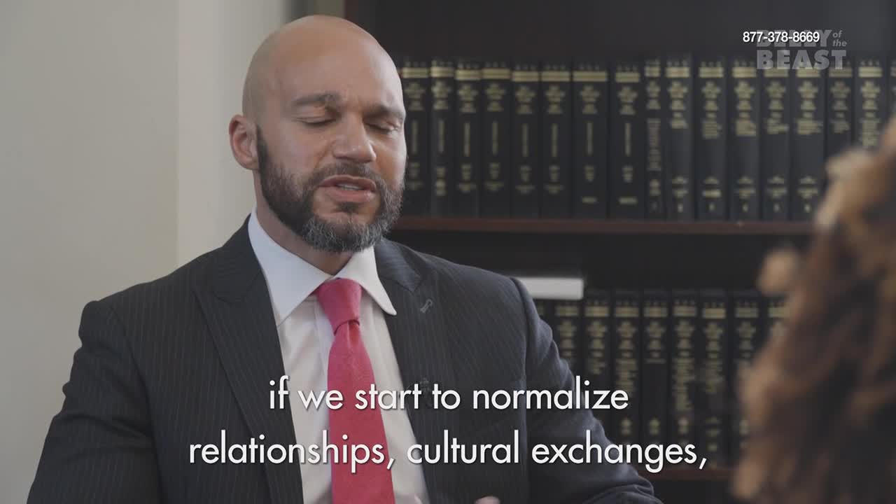 A man in a suit and pink tie speaks with his eyes closed, his brow furrowed. Behind him, rows of law books fill a shelf.