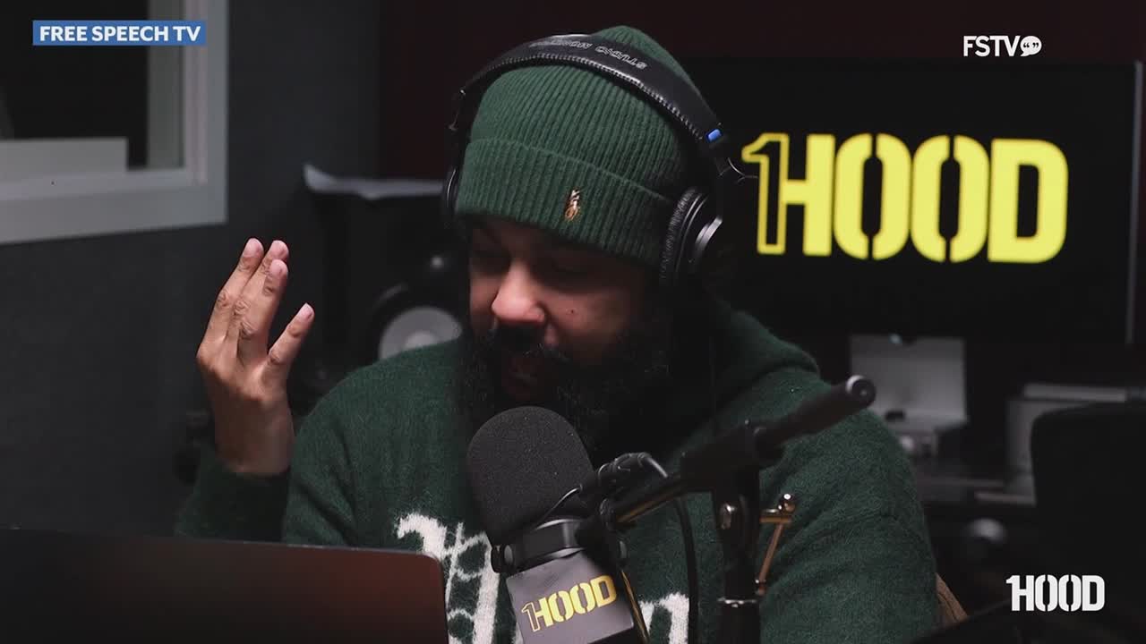 A man wearing a green beanie and headphones gestures with his left hand while speaking into a microphone. The "HOOD" logo appears on a screen behind him, broadcasting on Free Speech TV.