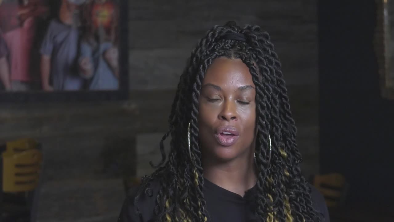 A woman with long, braided hair speaks directly to the camera. Behind her, a framed photograph of a family hangs on a textured wall.