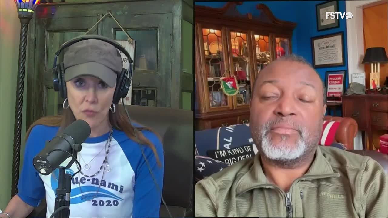 A woman wearing a baseball cap and headphones speaks into a microphone, her blue and white "Blue-nami 2020" shirt visible. To her right, a man with a gray beard looks directly at the camera, his eyes closed, seated in front of a bookshelf and a flag-adorned pillow.