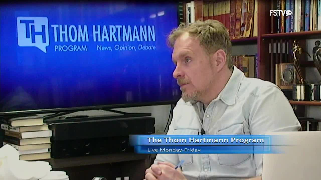 A man with a light blue shirt talks in front of a large screen displaying "The Thom Hartmann Program." The screen also shows "News, Opinion, Debate" and "Live Monday-Friday."