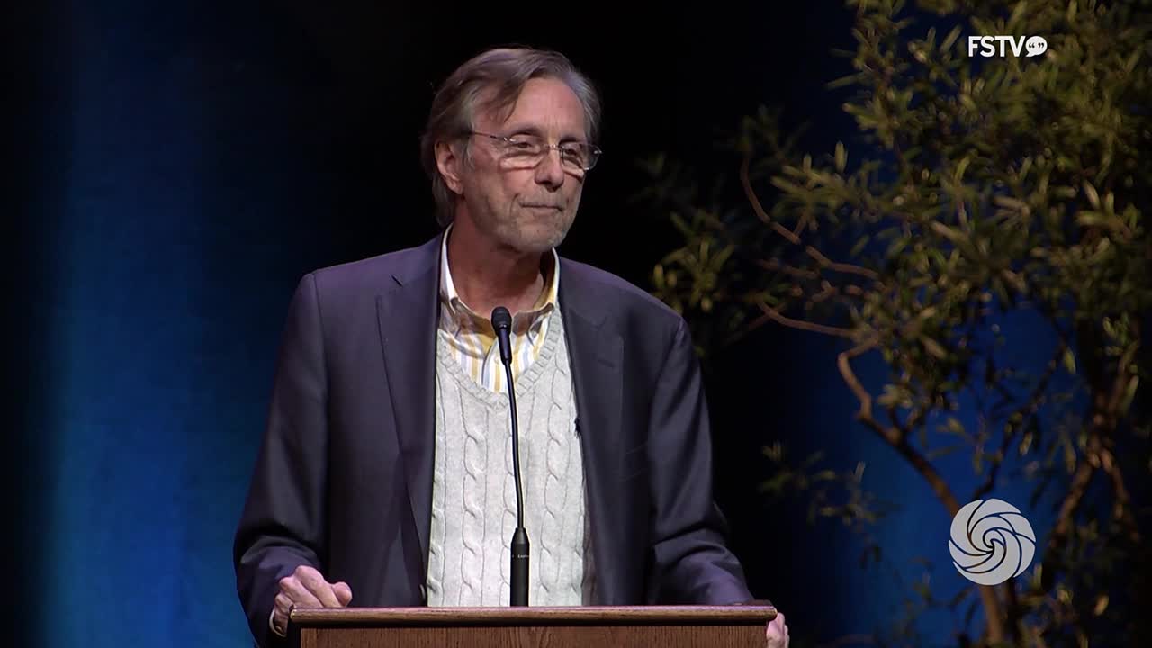 A man in a suit and sweater vest stands at a podium, speaking into a microphone. A logo for Free Speech TV appears in the corner of the screen.