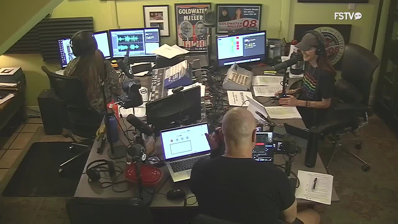 A woman in a baseball cap leans into a microphone, speaking into it. Across the desk, a man with a shaved head gestures with his hands while looking at a laptop. Posters of political candidates hang on the wall behind them.
